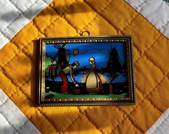 May include: A framed picture with a blue background depicting a couple standing in front of a windmill. The man is wearing a red hat and the woman is wearing a white cap. The picture is framed in gold.