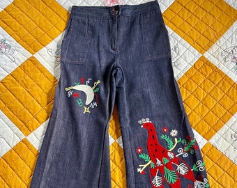 May include: Wide-leg denim jeans with embroidered designs. One leg features a red, green, and white bird with floral accents. The other leg features a crescent moon and stars in blue, red, and yellow.