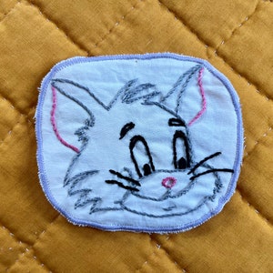 May include: A white fabric patch with an embroidered cartoon cat face. The cat has gray fur, black eyes, a pink nose, and black whiskers.