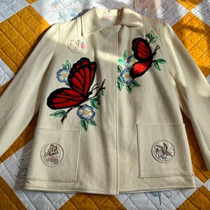 May include: A white wool jacket with embroidered butterflies and flowers. The jacket has two pockets and a button closure.