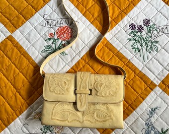 May include: A pale yellow leather crossbody bag with floral embossed detailing. The bag is laying on a yellow and white quilted fabric with embroidered flowers and state names.