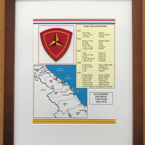 May include: Framed print featuring a red and yellow emblem, a map of Vietnam, and a list of battles from 1965-1970. The print is encased in a brown wooden frame with a white matting.