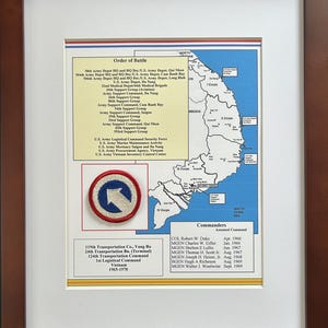 May include: Framed military memorabilia with a map of Vietnam and an Order of Battle list. Includes a patch with a blue arrow on a red and white background. The frame is a dark brown colour.