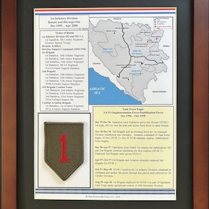 May include: A framed military display featuring a map of Bosnia and Herzegovina, an Order of Battle list for the 1st Infantry Division, a Task Force Eagle timeline, and an olive drab patch with a large red number one, all set against a black mat.