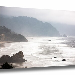 May include: A misty seascape with a rocky coastline and crashing waves. The water is a light blue and white, and the sky is a soft gray. The mountains in the background are covered in fog.