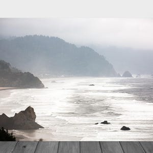 May include: A misty, foggy landscape with a rocky coastline and a vast ocean. The water is a light blue and white, with waves crashing on the shore. The sky is a light gray, with clouds obscuring the sun. The mountains in the background are a dark green.