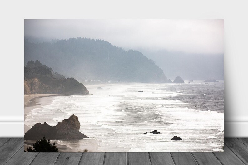 May include: A misty, foggy landscape with a rocky coastline and a vast ocean. The water is a light blue and white, with waves crashing on the shore. The sky is a light gray, with clouds obscuring the sun. The mountains in the background are a dark green.