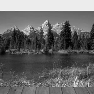 May include: A black and white photograph of a mountain range reflected in a still lake. The mountains are snow-capped and the trees in the foreground are silhouetted against the water.
