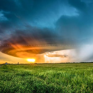 May include: A dramatic sunset with a large, dark cloud formation in the sky. The cloud is illuminated from below by the setting sun, creating a bright orange glow. The cloud is casting a shadow on the green grass field in the foreground.