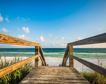 Peut inclure: Une passerelle en bois avec des rampes mène à une plage de sable blanc avec une eau bleu turquoise. Le ciel est bleu vif avec des nuages blancs.