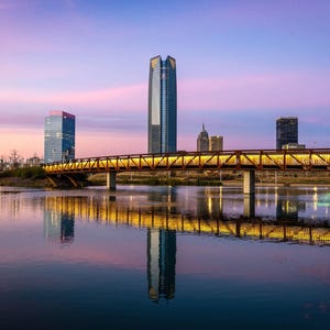 May include: A metal bridge over a body of water with a city skyline in the background. The sky is a vibrant pink and purple, and the water reflects the city lights.