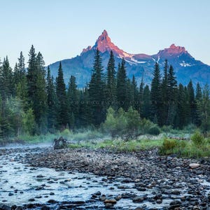 May include: A scenic view of a mountain range with a river flowing through the foreground. The mountains are covered in trees and the river is surrounded by rocks. The sky is a light blue color and the mountains are a dark blue color.