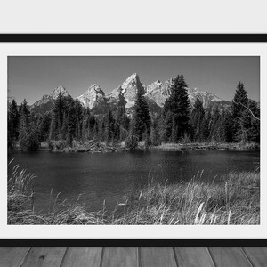 May include: Black and white photograph of a mountain range reflected in a still lake. The mountains are snow-capped and the trees in the foreground are silhouetted against the water.