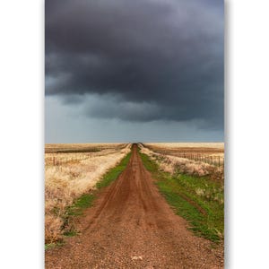 May include: A dirt road stretches into the distance, disappearing under a dark, ominous storm cloud. The road is flanked by dry, brown grass on either side.