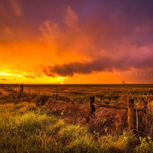 May include: A scenic sunset over a grassy field with a wooden fence in the foreground. The sky is a vibrant orange and purple, with clouds creating a dramatic effect.