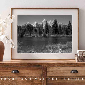 May include: Black and white photograph of a mountain range reflected in a lake. The mountains are tall and jagged, and the lake is calm and still. The photo is framed in a simple wooden frame.