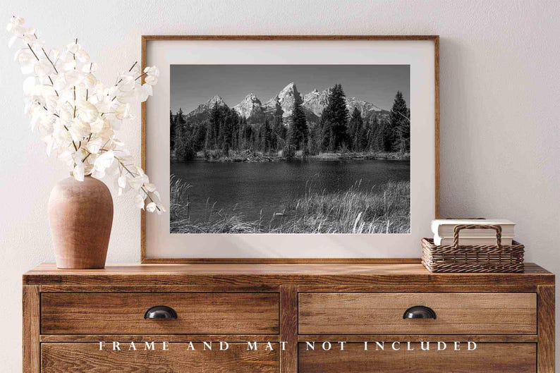 May include: Black and white photograph of a mountain range reflected in a lake. The mountains are tall and jagged, and the lake is calm and still. The photo is framed in a simple wooden frame.