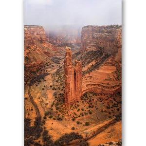 May include: Aerial view of a canyon landscape featuring a tall, reddish-brown rock formation in the center. The canyon walls are layered with similar hues, and a road winds through the valley. Overcast sky.