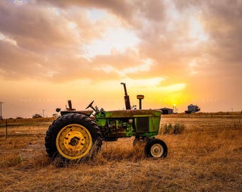 May include: A green and yellow tractor sits in a field at sunset. The tractor is facing away from the camera and is partially obscured by a cloud of dust.