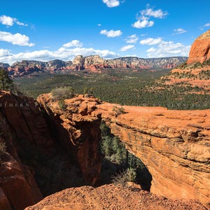 May include: A natural sandstone archway in Sedona, Arizona, with a view of the red rock formations and a blue sky with white clouds.