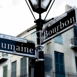 May include: Two black and white street signs with white lettering. The top sign reads "Rue Bourbon Bourbon" and the bottom sign reads "Rue Dumaine Dumaine".