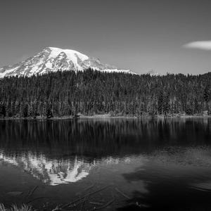 May include: A black and white photograph of a snow-capped mountain range reflected in a still lake. The mountain range is covered in trees and the lake is surrounded by trees.