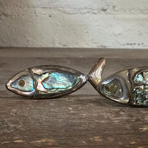 Puede incluir: Dos objetos decorativos con forma de pez hechos de metal con incrustaciones de segmentos de concha de abulón iridiscente. Cada pez presenta un detalle de ojo circular y un diseño de cuerpo segmentado en tonos brillantes azules, verdes y plateados.