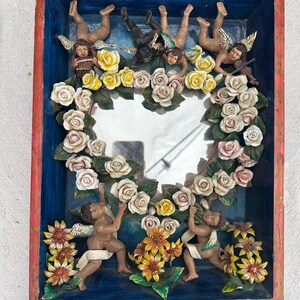 May include: A rectangular, hand-painted shadow box with a blue interior and red trim. The box features cherubic figures, a heart-shaped mirror, and floral arrangements of white, yellow, and orange flowers. The cherubs are in various poses.