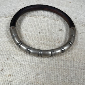 Op de afbeelding: Een zilveren en donkerbruine armband met een decoratief ontwerp. De armband is gemaakt van metaal en hout.