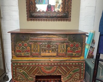 May include: A vintage wooden chest with intricate hand-painted designs in shades of green, red, yellow, and blue. The chest has a carved wooden top and sits on four carved legs.