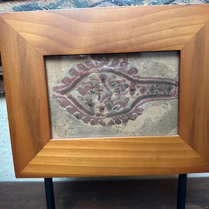 May include: A framed piece of antique fabric with a red, green, and blue paisley design. The fabric is displayed on a wooden stand.