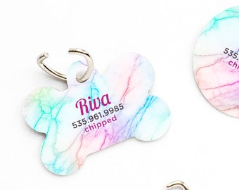 May include: Three colorful dog tags with a watercolor design. The tags are shaped like a bone, a circle, and a heart. Each tag has the name "Riva" and a phone number, along with the word "chipped".