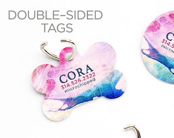 May include: Two double-sided dog tags with a pink, blue, and white watercolor design. The tags are shaped like a bone and a circle. The text on the tags reads "CORA 314.526.2322 microchipped".