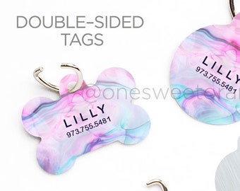 May include: Two double-sided dog tags with a pink and blue marble design. The tags are shaped like a bone and a circle. The text on the tags reads "LILLY" and "973.755.5481".