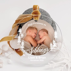 May include: Clear acrylic Christmas ornament with a photo of two babies, text that reads "First Christmas as Siblings", and the names "Liam & Brian" with "Christmas 2025". The ornament has a gold ribbon.