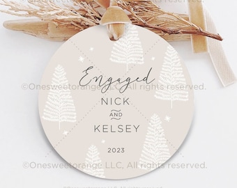 May include: A beige round ornament with a white stylized tree design and the text "Engaged Nick and Kelsey 2023".
