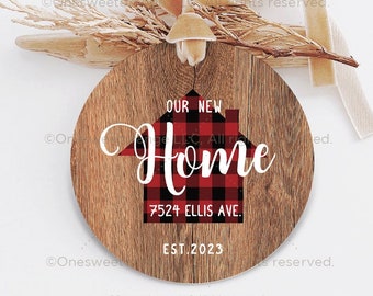 May include: Round wooden ornament with a red and black buffalo plaid house design. The text "Our New Home" is written in white cursive font. The address "7524 Ellis Ave." and the year "EST. 2023" are also written in white.