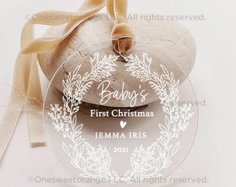 May include: A clear acrylic Christmas bauble with a white floral wreath design. The bauble features the text "Baby's First Christmas" with a heart and the name "JEMMA IRIS" and the year "2021".