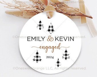 May include: A white round ornament with black and white plaid Christmas trees and the words "EMILY & KEVIN engaged 2024" in black and gold lettering. A beige ribbon and dried plant sprigs are attached.