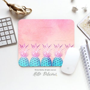 May include: A rectangular mousepad with a pink watercolor background and a row of stylized pineapples in shades of blue and purple. A white computer mouse, keyboard, and other office supplies are arranged around the mousepad.