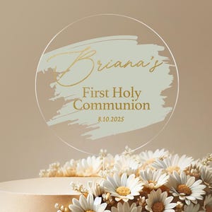 May include: A round, clear acrylic cake topper with a light green brushstroke design and gold lettering. The text reads "Briana's First Holy Communion 8.10.2025".