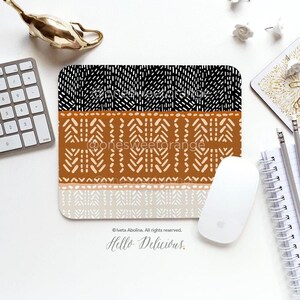 May include: A brown, black and white patterned mouse mat with the text "Hello Delicious." The mouse mat is on a white surface with a gold metal decorative object, a keyboard, a white mouse and a notebook.