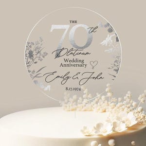 May include: A clear, circular cake topper with silver text celebrating a 70th Platinum Wedding Anniversary. The design includes floral accents and the names "Emily & John" with the date 8.17.1974. The topper is placed on a white cake decorated with white flowers.