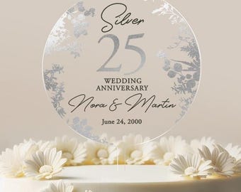 May include: Silver 25th wedding anniversary cake topper with a floral design. The topper is round and features the text "Silver 25 Wedding Anniversary Nora & Martin June 24, 2000".