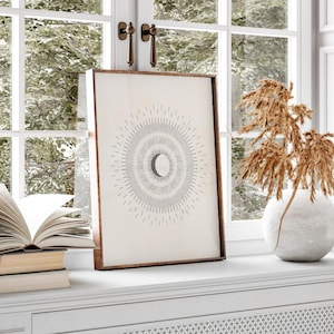 May include: A minimalist black and white print of a crescent moon inside a circle of radiating lines. The print is framed in a light brown wood frame and is displayed on a white shelf.