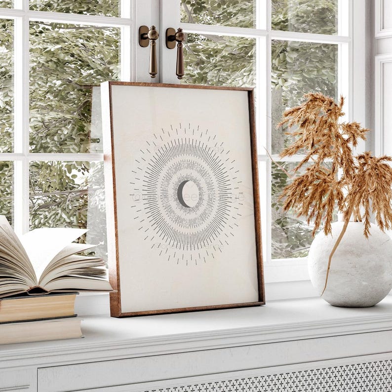 May include: A minimalist black and white print of a crescent moon inside a circle of radiating lines. The print is framed in a light brown wood frame and is displayed on a white shelf.
