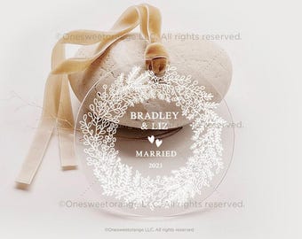 Könnte beinhalten: Ein runder, transparenter Weihnachtsbaumschmuck mit einem weißen floralen Kranzdesign. Der Schmuck hat den Text "Bradley & Liz Married 2021" innerhalb des Kranzes.