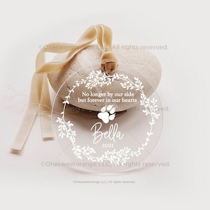 May include: A clear round ornament with a white floral wreath, a paw print, and the text "No longer by our side but forever in our hearts Bella 2021".