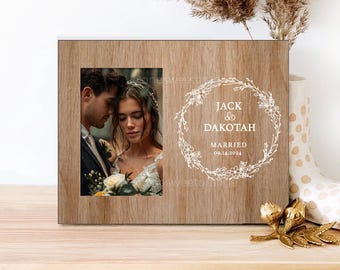 May include: A wooden picture frame with a photo of a couple in front of a floral wreath design. The wreath has the text "Jack & Dakotah Married 09.14.2024".