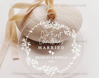Könnte beinhalten: Ein kreisförmiges Ornament aus klarem Acryl mit einem weiß aufgedruckten botanischen Kranzmotiv. Der Text lautet Our First Christmas MARRIED, BRADLEY & KENDAL und 2021, ergänzt durch zwei kleine Herzen. Ein beiges Samtband ist oben durchgezogen.